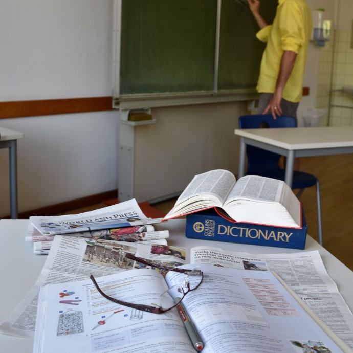 Das Bild zeigt einen Tisch mit Lernutensilien für den Sprachunterricht und dem Lehrer an der Tafel. 