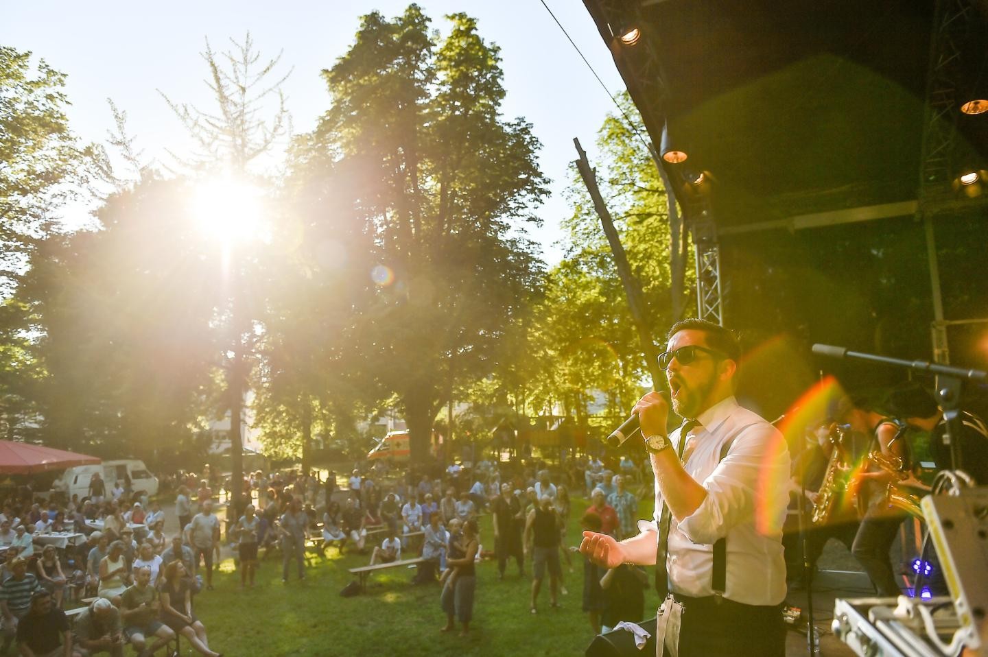 Ein Mann mit Sonnenbrille und weißem Hemd steht auf einer Bühne bei einem Outdoor-Festival, hält ein Mikrofon in der Hand und spricht enthusiastisch in die Menge. Im Hintergrund scheint die Sonne durch hohe Bäume, beleuchtet Zelte, eine jubelnde Zuschauermenge auf dem Gras und Bühnenbeleuchtung, was eine lebendige, sommerliche Atmosphäre erzeugt.