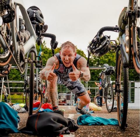 Triathlon Radzone 2 Ein tätowierter Triathlet jubelt mit beiden Daumen nach oben in einer Wechselzone zwischen Rennrädern.