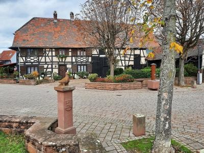 Das Bild zeigt den Dorfplatz in Bruchhausen mit einem Fachwerkhaus im Hintergrund. Im Vordergrund sind gepflasterte Wege, Bäume und Skulpturen zu sehen. Dorfplatz Bruchhausen