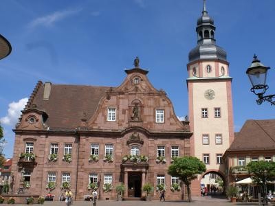 Das Bild zeigt das historische Rathaus von Ettlingen mit seinem markanten Rathausturm vor einem blauen Himmel. Der Platz davor ist belebt und von Pflanzen und Straßenlaternen gesäumt. Ettlinger Rathaus mit Rathausturm