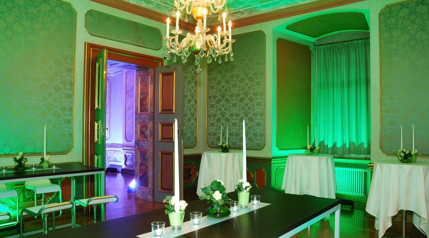 Grüner Saal mit grünem Licht und Stehtischen auf denen weiße Kerzen und Blumen stehen