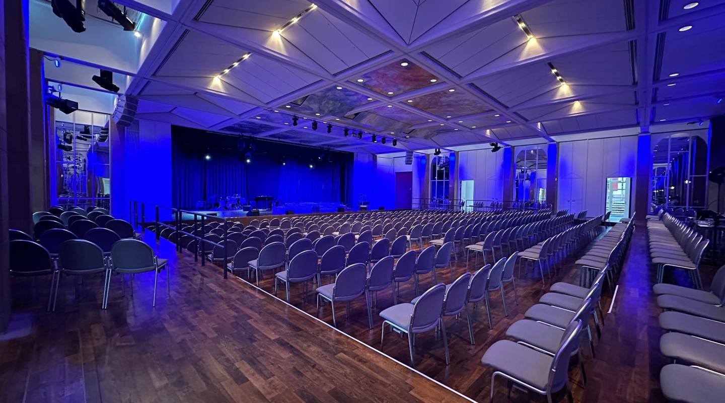 Schlossgartenhalle von innen mit roter und blauer Beleuchtung an den Wänden, Reihenbestuhlung mit Gästen und einer Sängerin sowie einem Sänger auf der Bühne neben dem Flügel