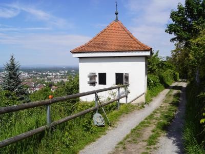 Weißes Häusle auf dem Panoramaweg Ein kleines, weißes Gebäude mit einem roten Ziegeldach am Rande eines schmalen Schotterwegs. Der Weg ist von einem Holzgeländer und grüner Vegetation gesäumt. Im Hintergrund erstreckt sich eine Stadtlandschaft unter einem blauen Himmel. Bildunterschrift: "Weißes Häusle Panoramaweg".