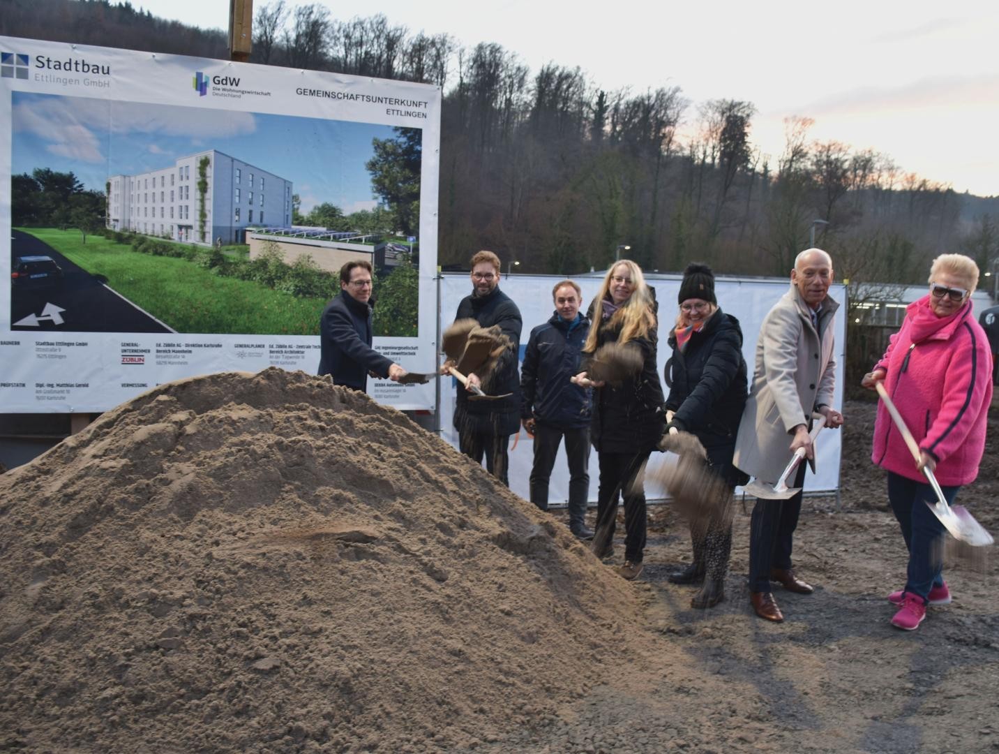 Erster Spatenstich für das Bauprojekt: Sieben Personen schaufeln vor der Infotafel den Sand, es sind Vertreter des Aufsichtsrates, der Geschäftsführer der Stadtbau und ein Vertreter der Baufirma.
