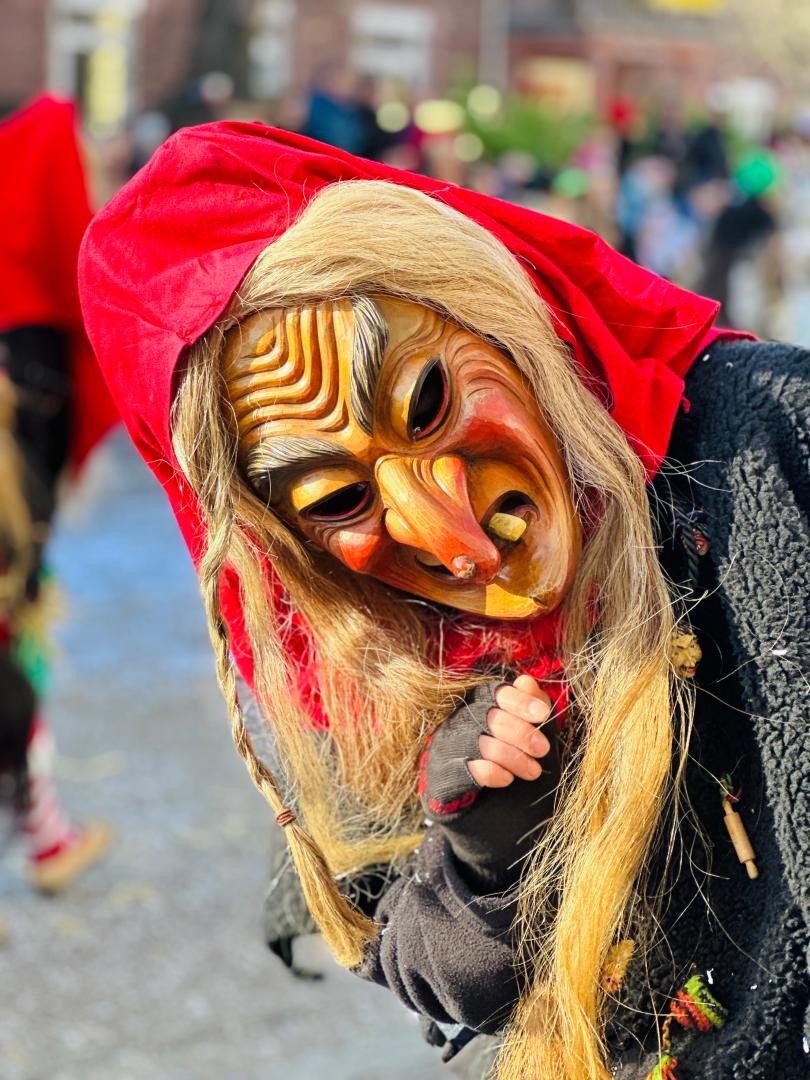 Narr in einem Hexenkostüme mit einem roten Kopftuch welcher sich zur seite neigt.