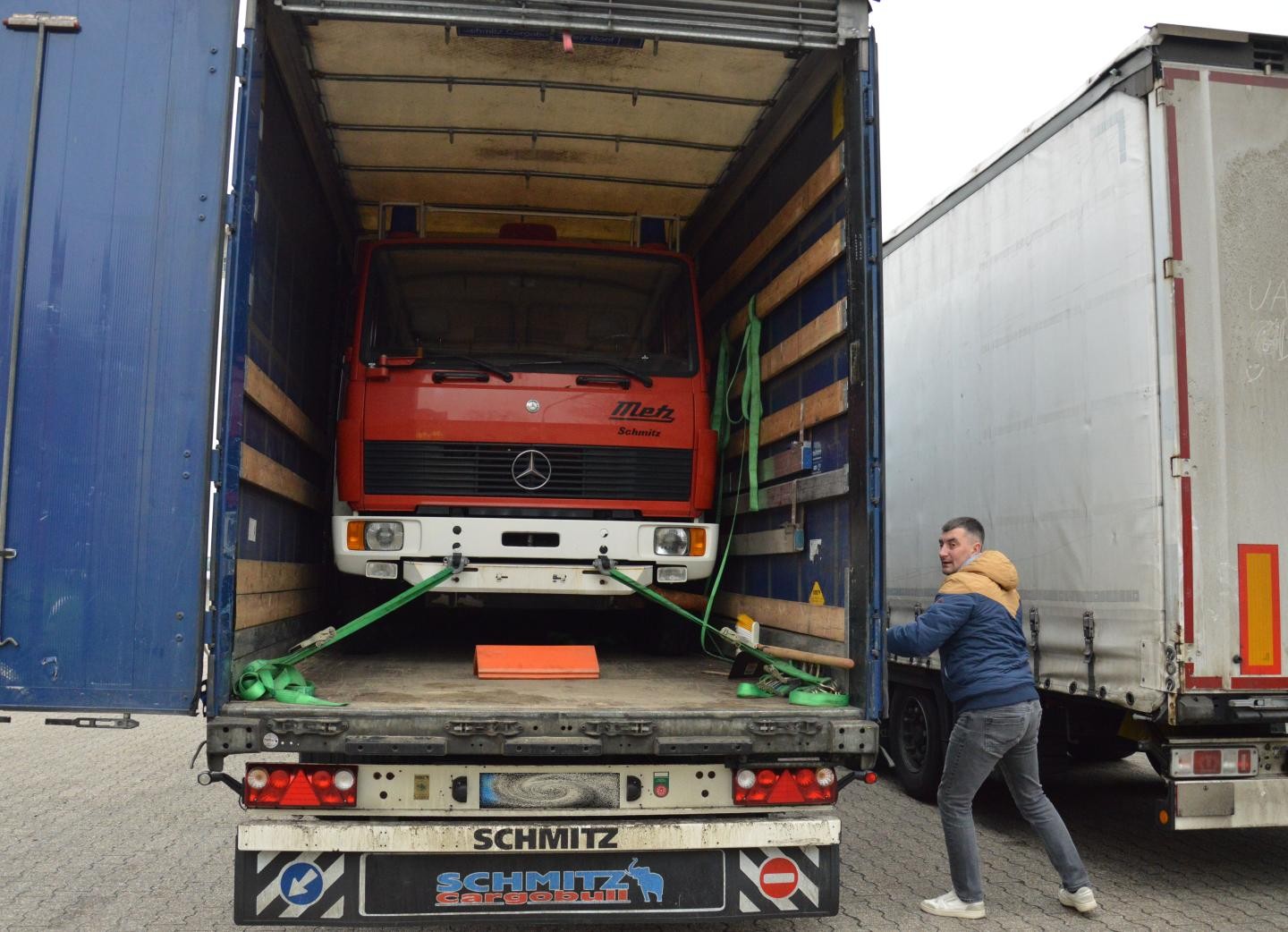 Die hintere Tür eines großen LKWs wird vom Fahrer geöffnet und man sieht im Innern das Feuerwehrfahrzeug. 