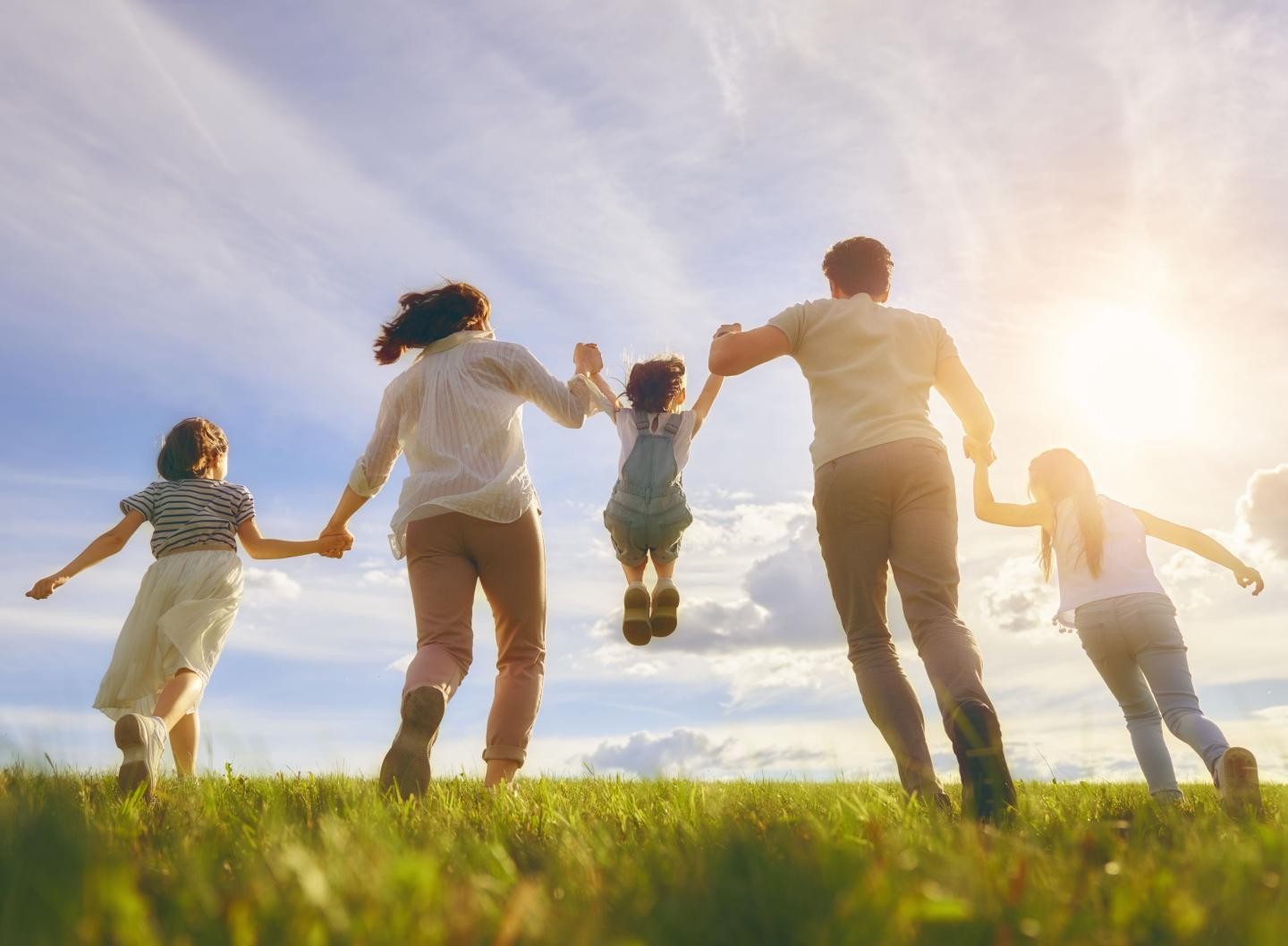 Eine fünfköpfige Familie in Rückansicht, alle halten sich an den Händen und sie laufen über eine Wiese, die Sonne scheint. 