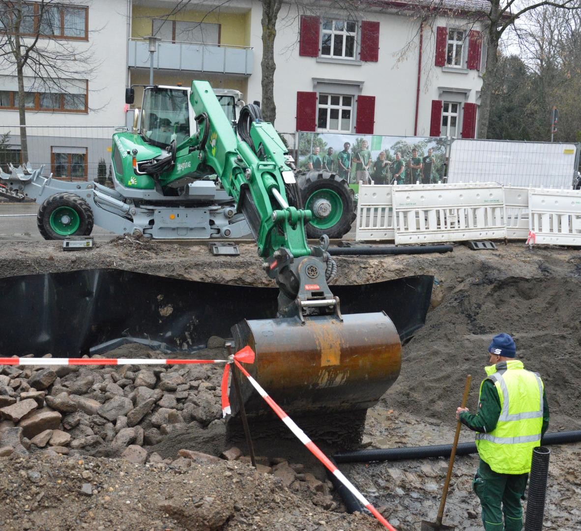 Eine Baggerschaufel schüttet Baumsbustrat in die künftige Baumgrube, davor steht ein Mitarbeiter, der das Substrat verteilen wird. 