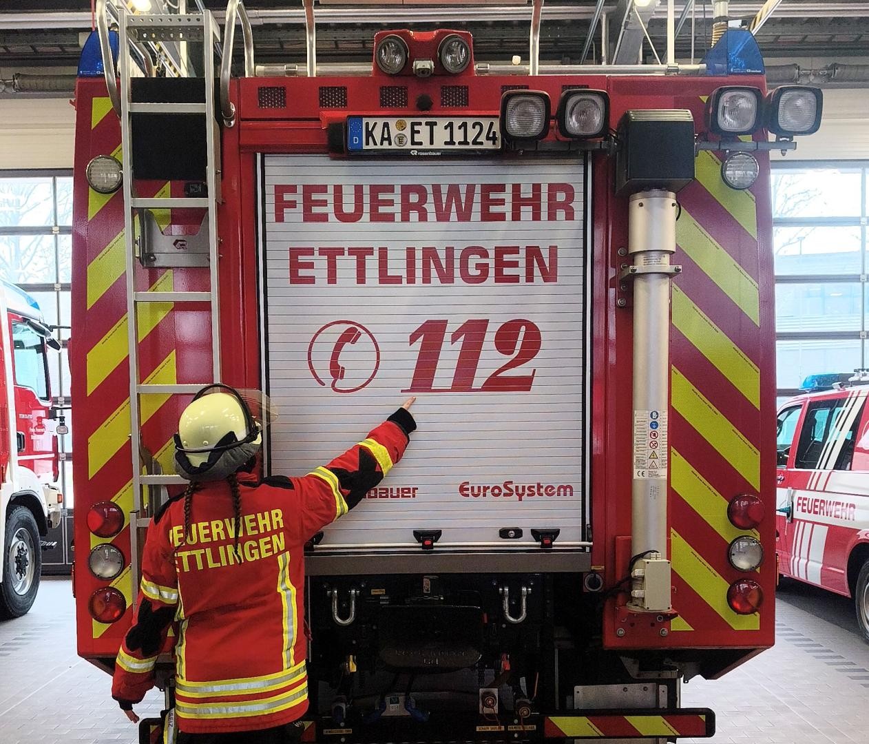 Ein Feuerwehrmitglied zeigt auf der Fahrzeugrückseite auf die dort aufgedruckte Notrufnummer 112