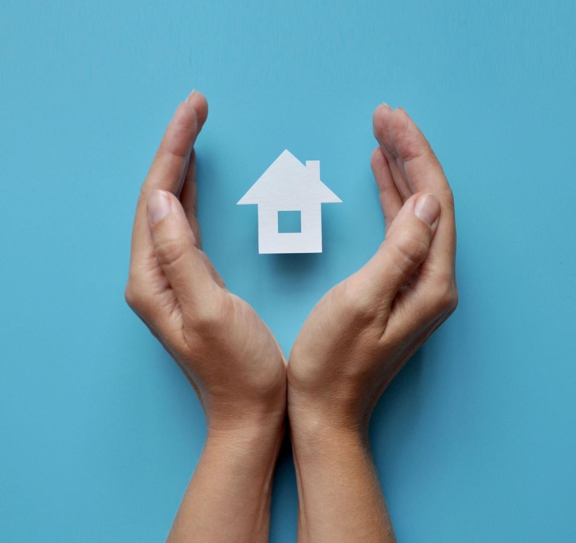 zwei Händen bilden vor blauem Hintergrund ein Oval, in dem symbolhaft ein kleines Häuschen steht. 
