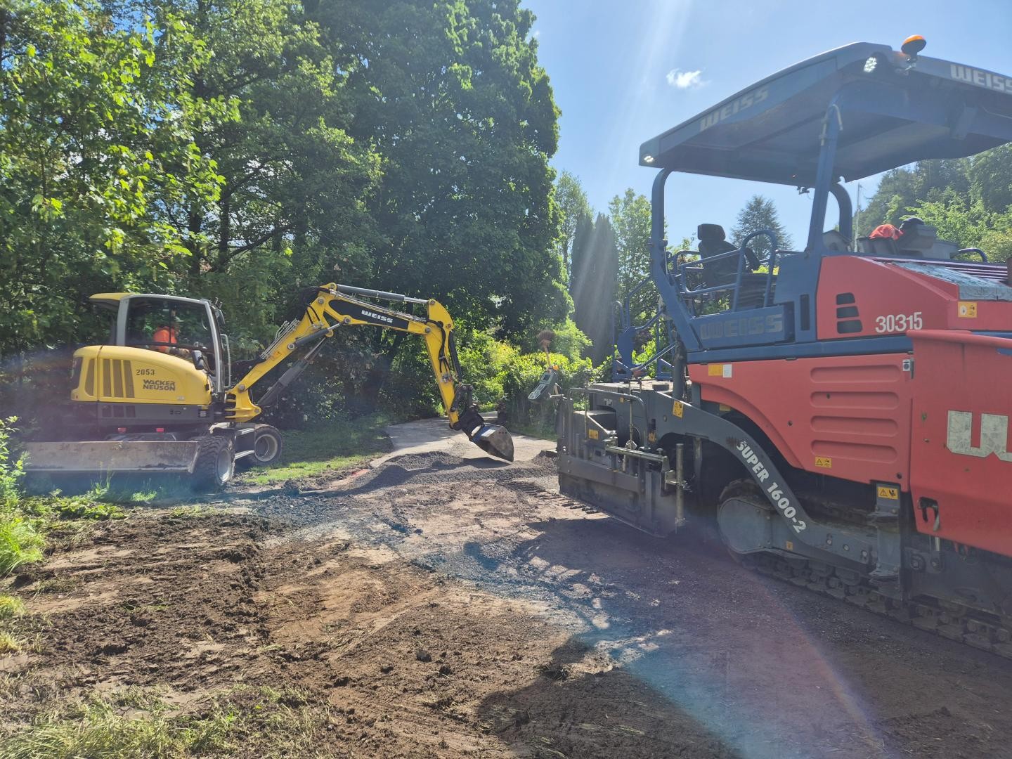 Graf-Rhena-Weg Ein Bagger schaufelt die neue Deckschicht auf den Weg und ein Planierer fährt anschließend darüber