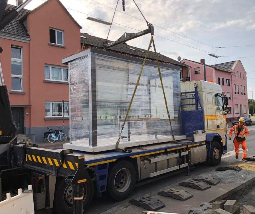 Auf einem Tieflader wird ein Wartehäuschen angeliefert, das dann mit einem Kran an seinen Bestimmungsort gehoben wird. 