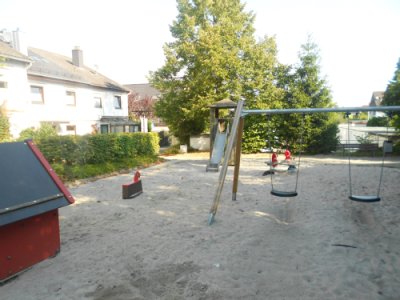 Schaukel mit zwei Sitzen auf Sandspielfläche sowie Hütte für Kleinkinder. Im Hintergrund Wipptiere und Rutschturm.